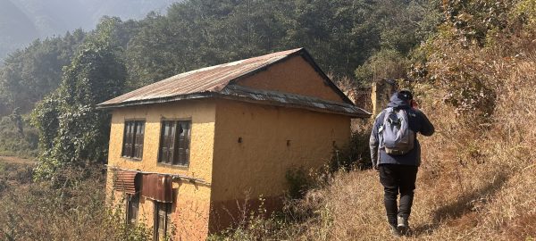 A trekker walking on a beginner-friendly trek in Nepal, passing by a traditional Nepali house painted yellow with a zinc roof.