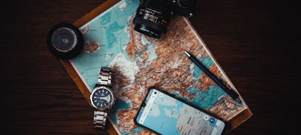 Top view of travel essentials including a map, camera, phone, watch, and lens on a wooden surface.